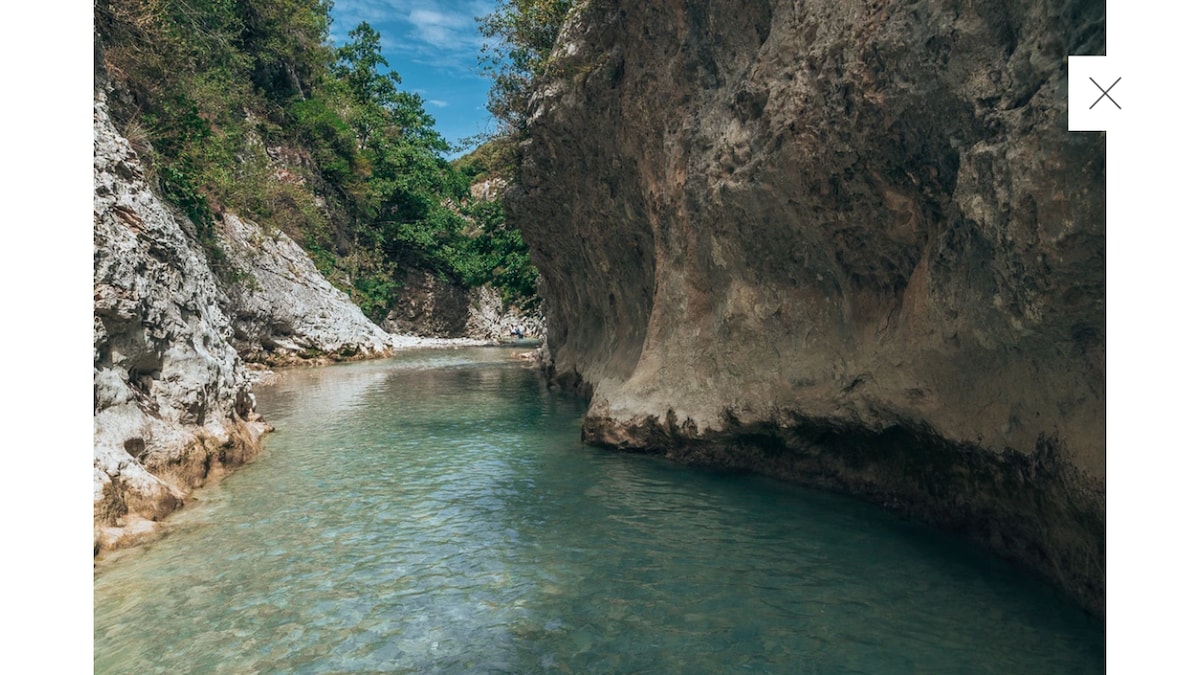 A tranquil waterway is surrounded by rocky cliffs and lush greenery. The clear water reflects the natural formations, creating a serene environment. Sunlight filters through the foliage, enhancing the blue-green hues of the water while shadows dance along the rocky surfaces.