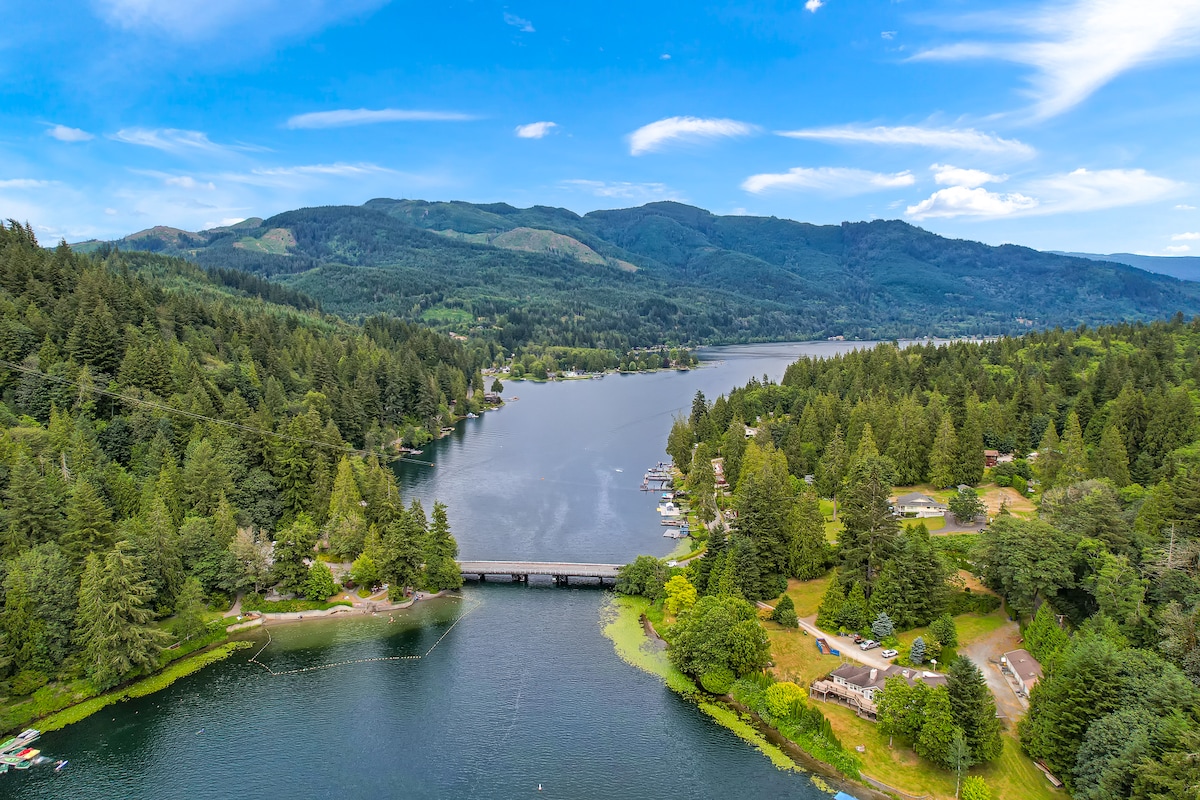 A tranquil aerial view captures the expansive Lake Samish surrounded by lush green forests and rolling mountains. The clear blue water reflects the sky, while a bridge spans across the lake, connecting both shores adorned with greenery and scattered homes.