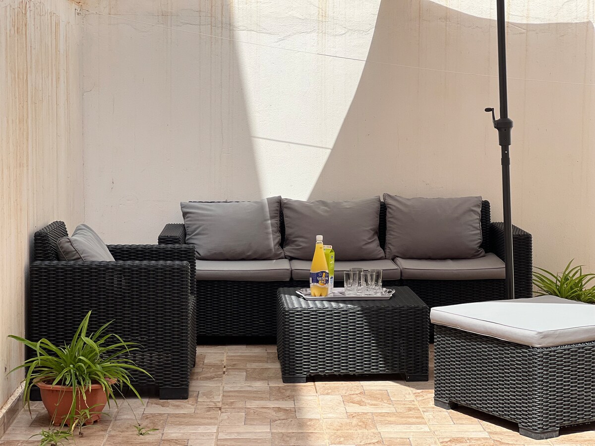 A cozy outdoor seating area is presented, featuring a dark wicker sofa with plush grey cushions. A glass coffee table sits in the center, accompanied by a pitcher and glasses. Potted plants add a touch of greenery, enhancing the inviting space.