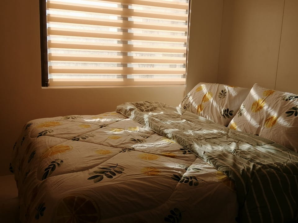 A cozy sleeping area features a spacious bed with a floral-patterned comforter and plush pillows. Warm light filters through the striped window blinds, creating a calming ambiance that highlights the inviting textures of the bedding.