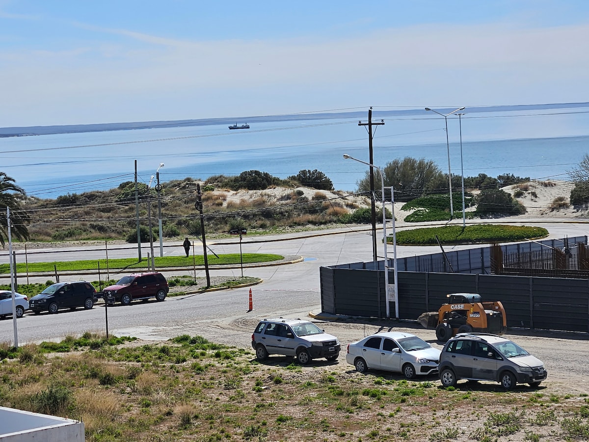A scenic view captures a quiet street leading to a beach. Vehicles are parked along the roadside, while a gentle sea sparkles in the background. The coastline's natural landscape is visible, complementing the tranquil atmosphere of the area.