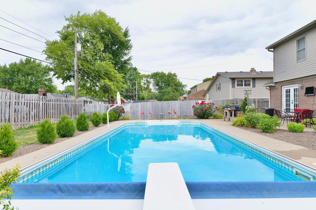 A private outdoor pool glistens under a cloudy sky, surrounded by well-maintained landscaping. A spacious deck features lounge chairs and a dining area, while a wooden fence offers privacy. Decorative flowers add a pop of color, creating a serene and inviting space.