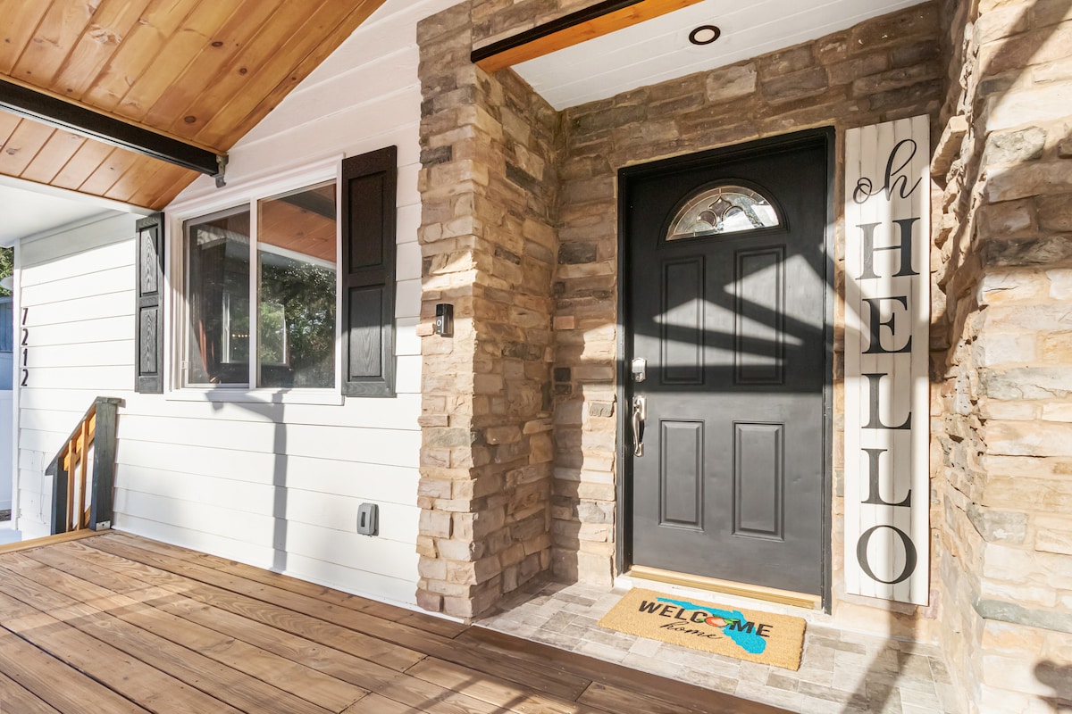 The entryway features a welcoming front door framed by natural stone accents and wooden beams overhead. A doormat with a friendly message is positioned on the ground. Large windows allow natural light to brighten the space.
