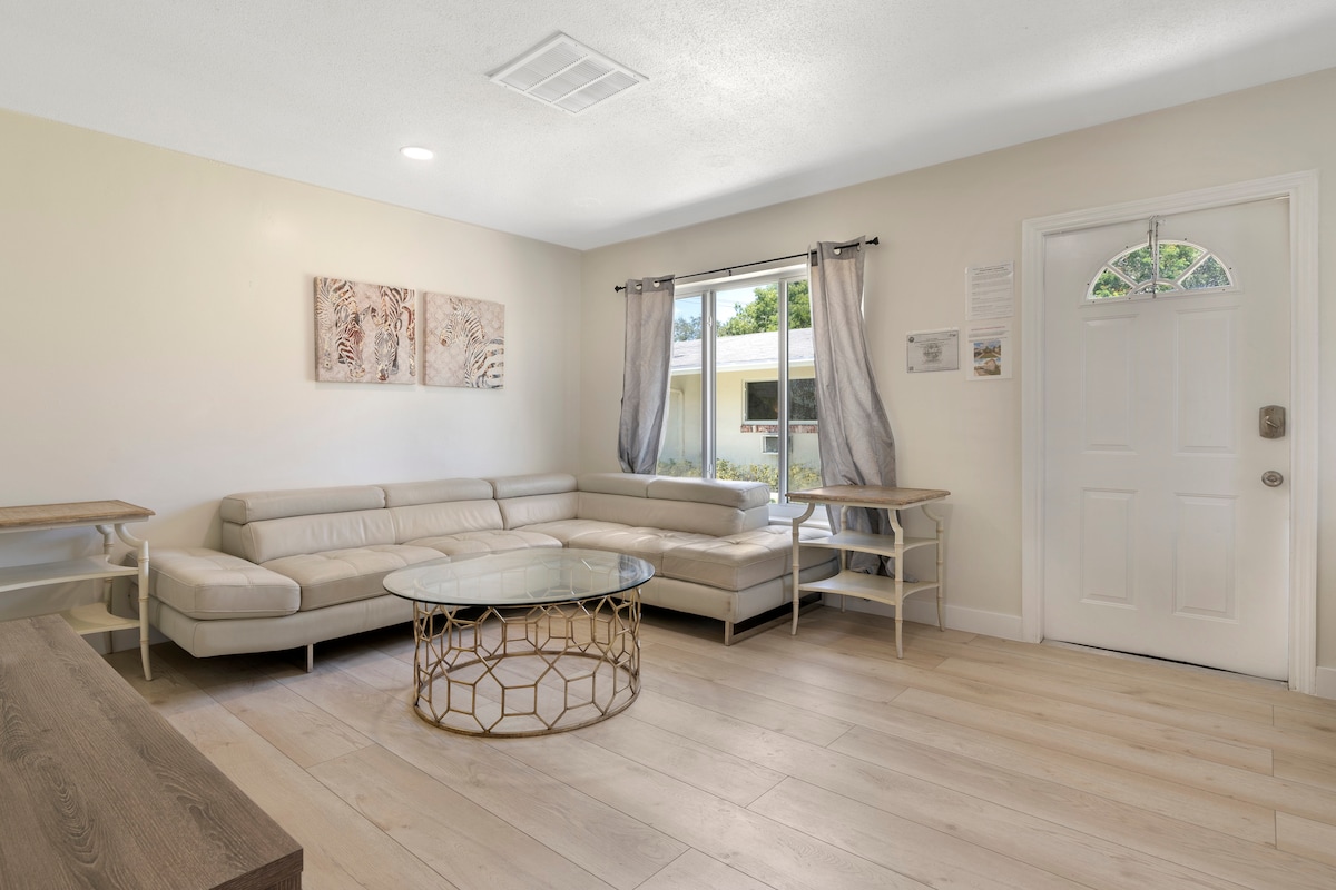 The living area features a modern sectional sofa arranged around a circular glass coffee table. Light-colored walls and a large window provide natural illumination, while soft curtains frame the view. Decorative art pieces and light wood flooring enhance the space's inviting layout.