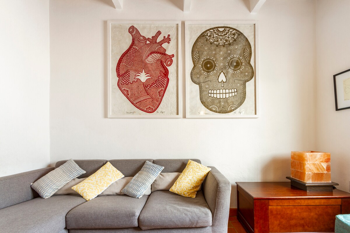 A cozy living area features a comfortable gray sofa adorned with decorative pillows in various patterns. Two artistic wall pieces, one depicting a heart and the other a mirror, are displayed above the sofa. A wooden side table and a light source add to the ambiance.