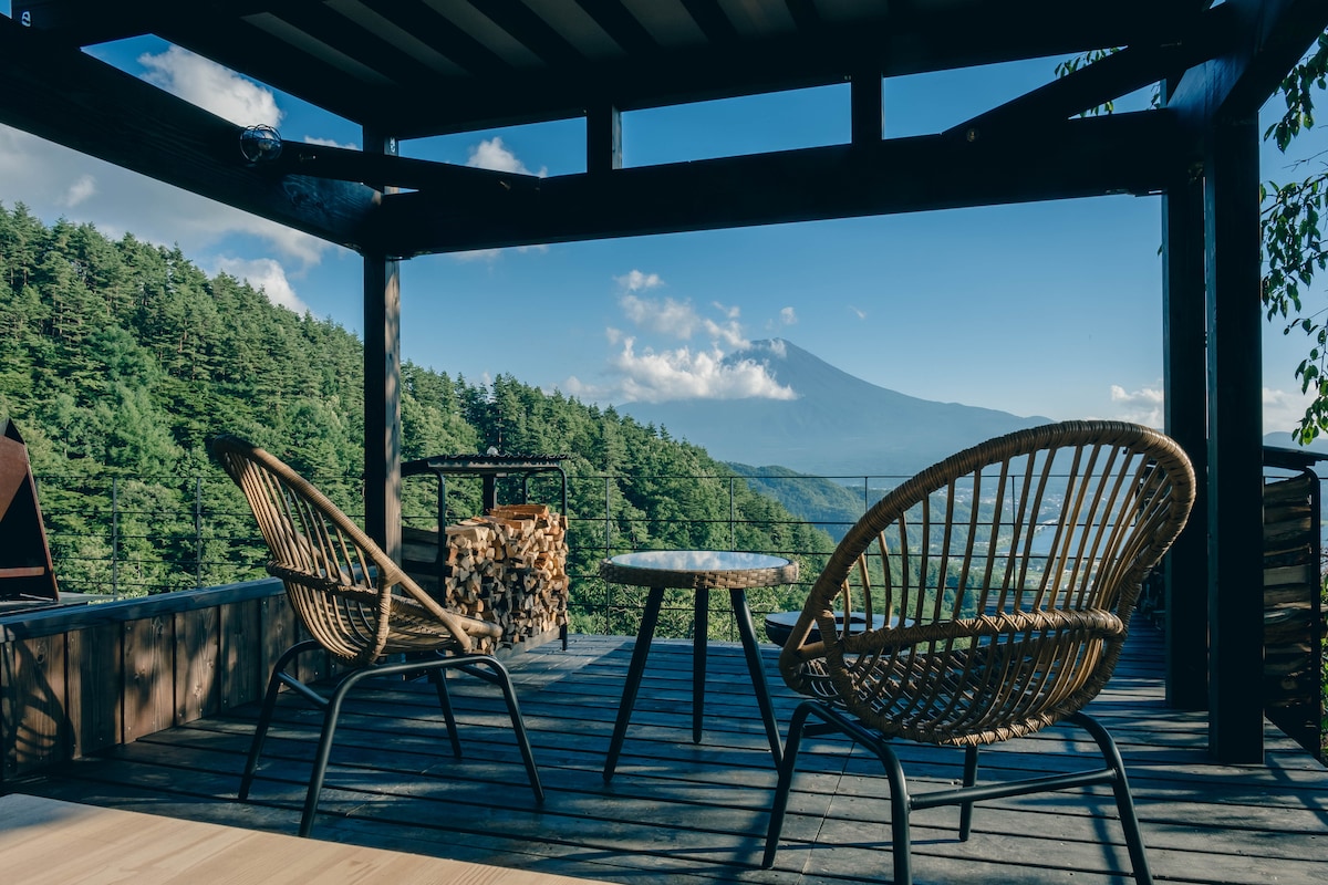 A spacious deck is presented with two comfortable, woven chairs and a small table, overlooking a scenic view of Mount Fuji. A stacked firewood display adds to the outdoor ambiance, framed by lush greenery and a clear blue sky.