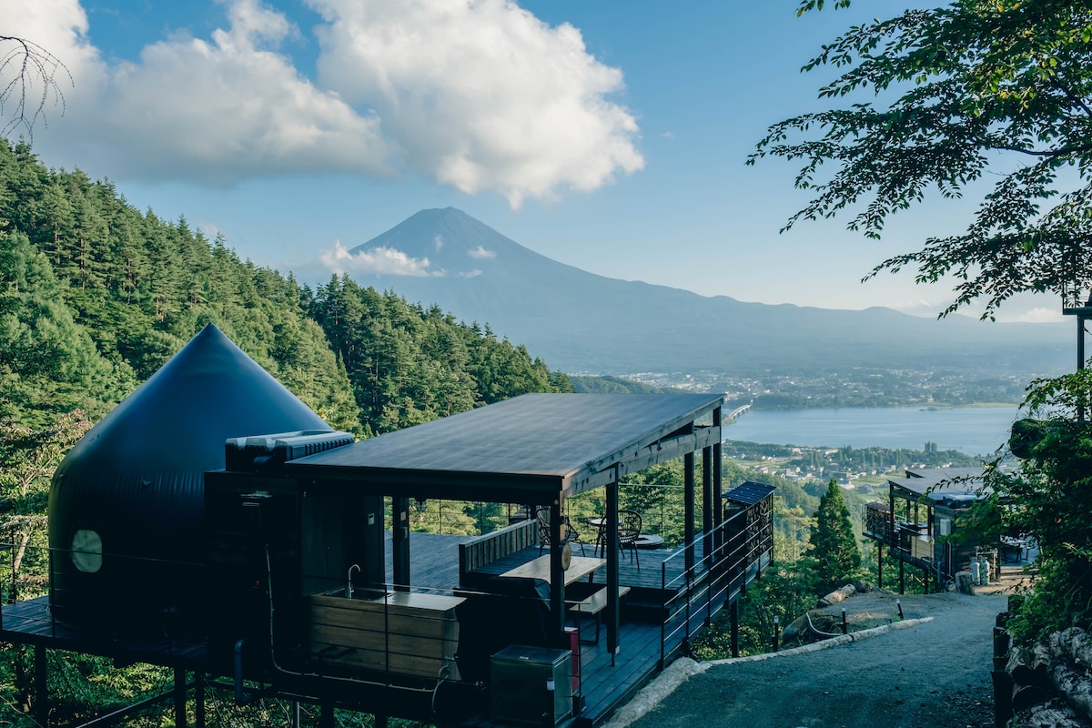 A modern accommodation structure is positioned on a mountainside, featuring a sleek black design. The scene includes lush greenery, with a clear view of Mount Fuji in the distance and Lake Kawaguchi below. Soft clouds drift across a blue sky, enhancing the tranquil atmosphere.