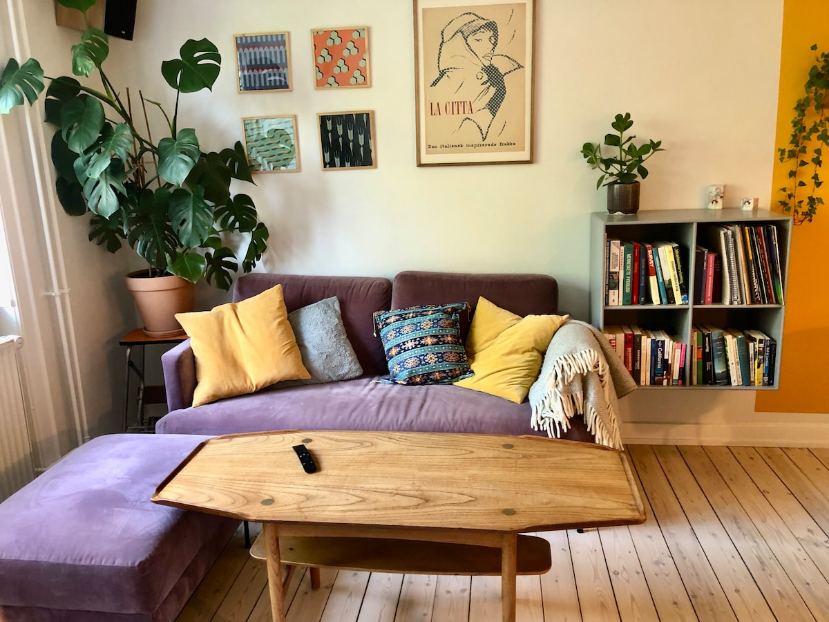 A purple sofa is positioned beside a small wooden coffee table, with a remote control resting on its surface. Colorful cushions lend comfort, while plants are visible near the furniture. Wall art and a bookshelf filled with books add character to the welcoming living area.