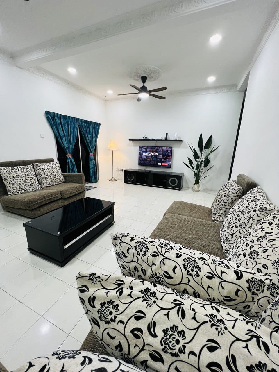 A spacious living area features a comfortable arrangement of sofas with floral-patterned cushions. A sleek coffee table sits at the center, while a wall-mounted television is visible above a low media console. Soft lighting is provided by a standing lamp, enhancing the inviting atmosphere.