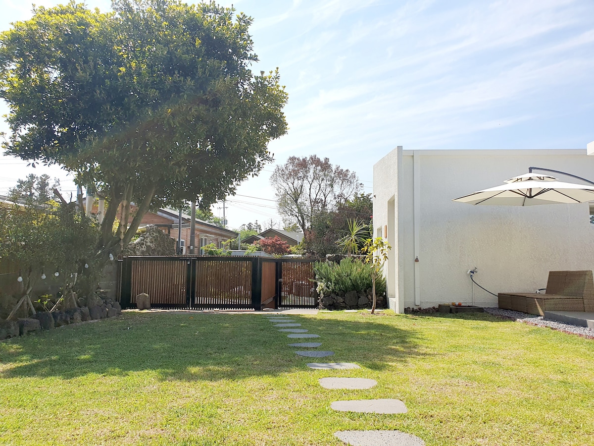 A neatly landscaped grassy yard features stone pathways leading toward a private entrance. A white structure is visible on one side, complemented by a large tree providing shade. An umbrella stands open, suggesting a comfortable outdoor space for relaxation.