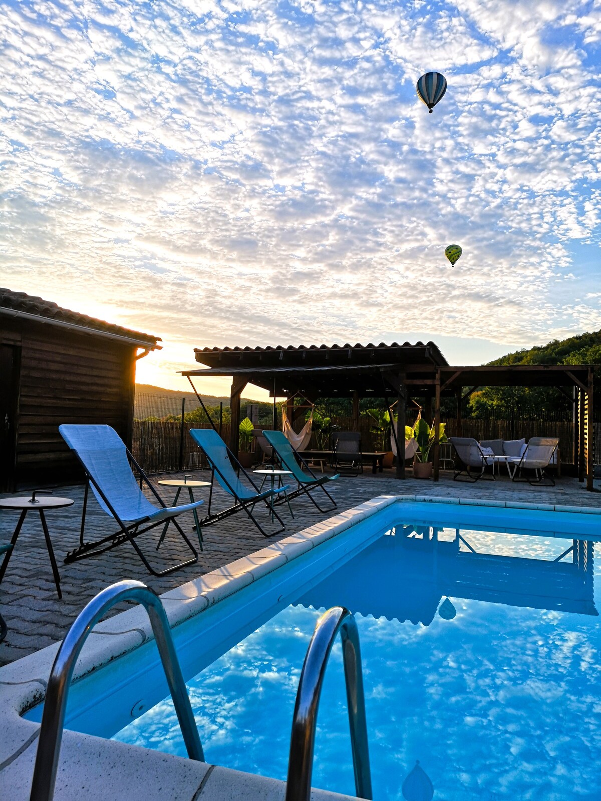 A serene swimming pool reflects the sky, framed by wooden decking and lounge chairs. A shaded seating area is visible nearby, surrounded by greenery. Hot air balloons can be seen in the distance against a beautiful sky, enhancing the tranquil ambiance of the outdoor space.