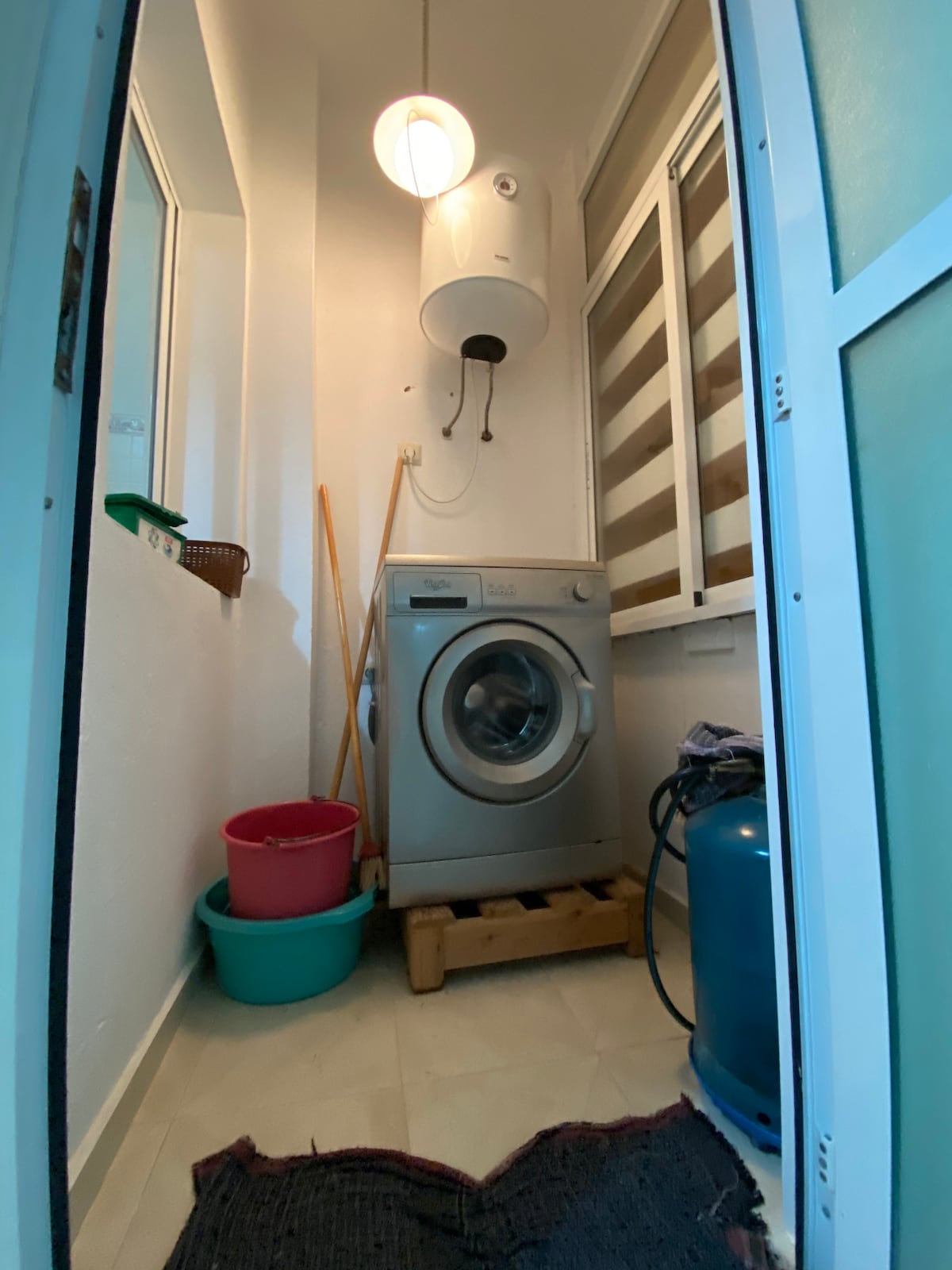 A utility area is seen featuring a washing machine placed on a wooden stand. Blue and pink containers are positioned nearby, along with brooms. Natural light filters in through a window, highlighting the clean, white walls and the simple layout of the space.