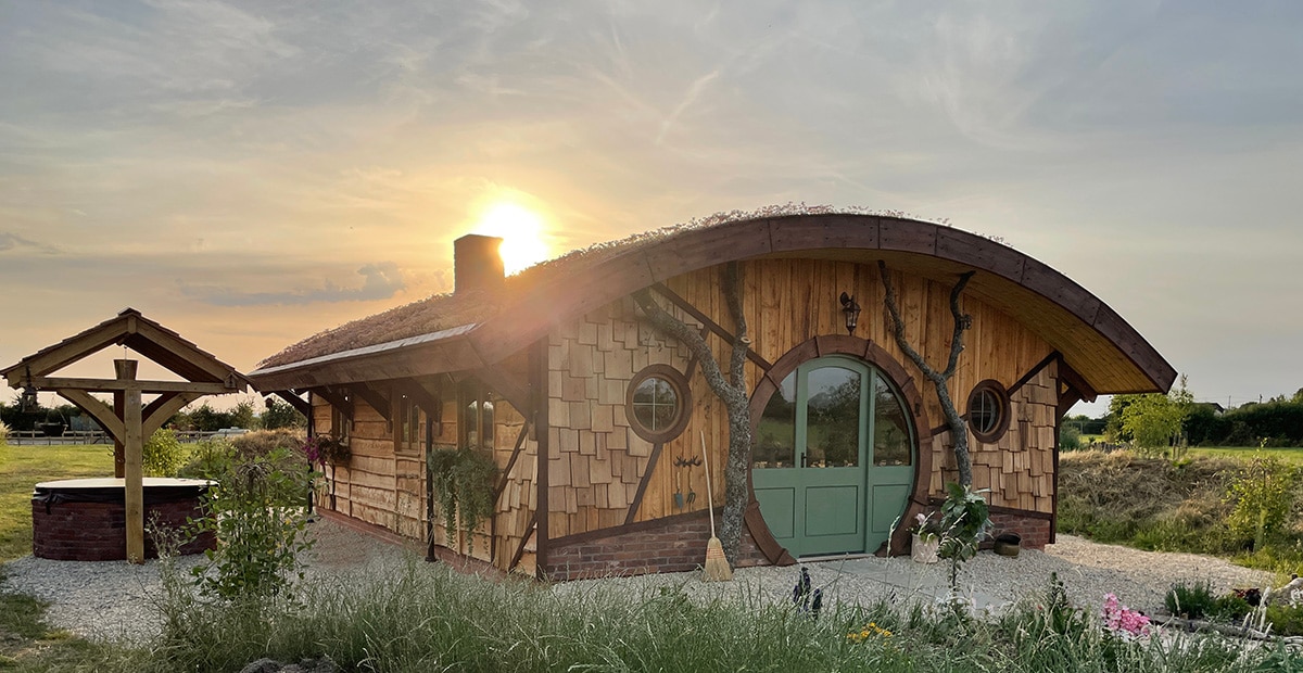 The exterior features a charming round structure with a wooden facade, accentuated by a large green door and a thatched roof. Sunlight casts a warm glow over the building, with a covered seating area nearby and verdant greenery surrounding the cottage.