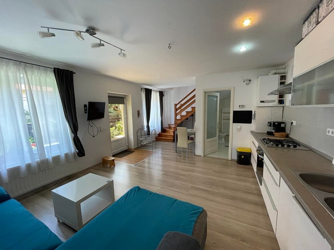 An open living area is visible, featuring a light-colored sofa and a coffee table. Large windows allow natural light to fill the space. A staircase leads to the upper level, and kitchen appliances are neatly arranged along the wall. Curtains frame the windows, enhancing the airy feel.