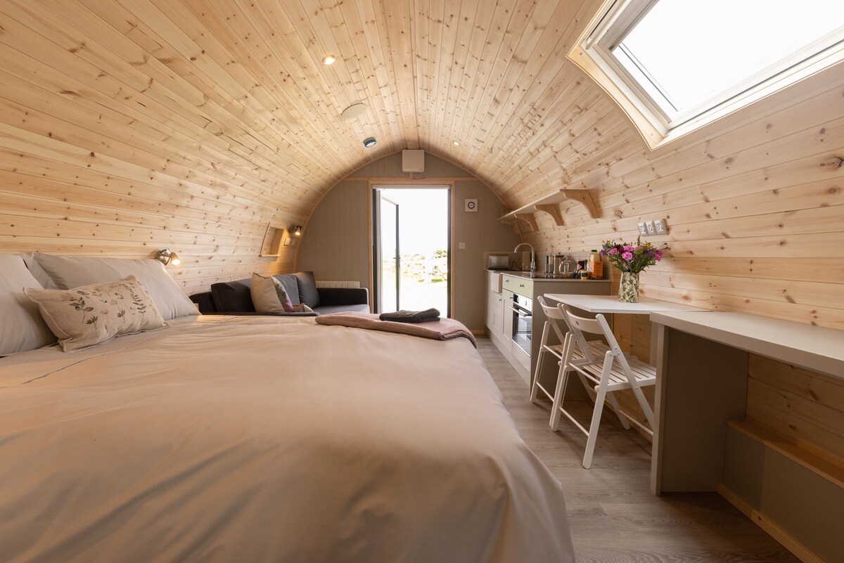 An interior glamping pod features smooth wooden paneling and a gently arched ceiling. A spacious bed is positioned alongside a seating area with a sofa. A small dining table with two chairs and a kitchenette are visible. Natural light floods through a skylight and the open door.