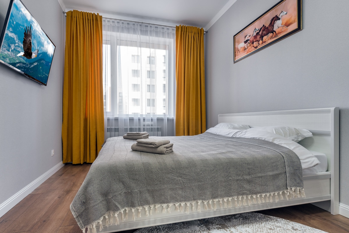 A spacious bedroom features a comfortable bed with light-colored linens and a decorative throw blanket. Two neatly arranged towels sit on the bed. Large windows are dressed with bright yellow curtains, and a flat-screen TV is mounted on the wall.