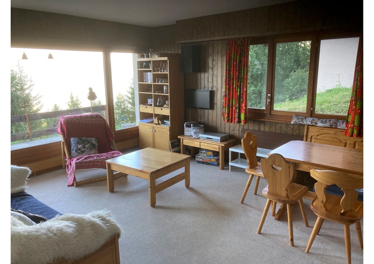 A spacious living area features large windows that invite natural light, offering views of the surrounding greenery. Wooden furniture is complemented by a cozy chair, a coffee table, and a dining table with carved chairs. Decorative curtains add a pop of color to the neutral decor.