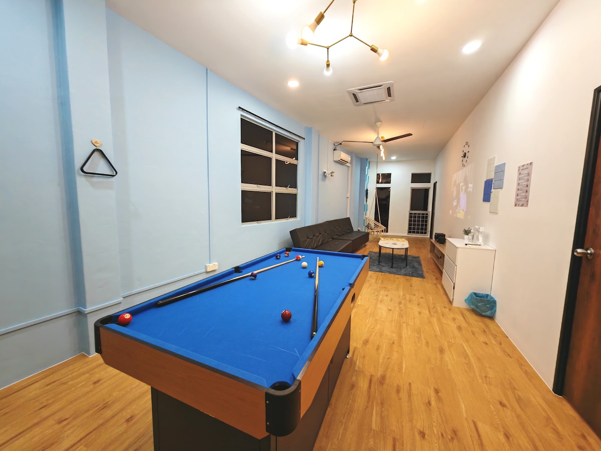 A spacious living area is highlighted by a blue snooker pool table, with colorful balls arranged on its surface. A sofa and coffee table are positioned nearby, while large windows allow natural light to fill the room, complemented by light-colored walls and wooden flooring.