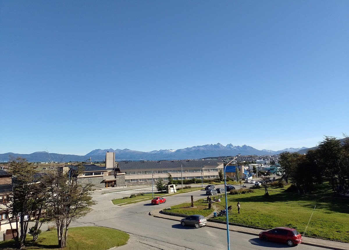 A panoramic view captures the bay of Ushuaia and mountain ranges under a clear blue sky. The foreground features lush green grass, parked vehicles, and a winding road that leads toward the waterfront, providing a sense of the surrounding urban setting.