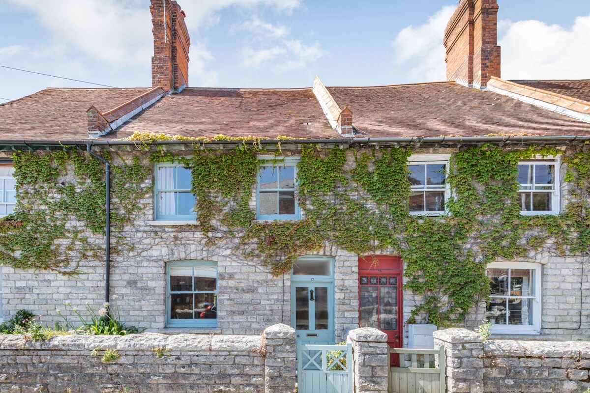 The charming exterior of a Victorian cottage is shown, with natural Purbeck stone facade and ivy covering the walls. Two large windows flank a central front door, accented by a light blue doorframe. A low stone wall is visible in the foreground, enhancing the inviting entrance.