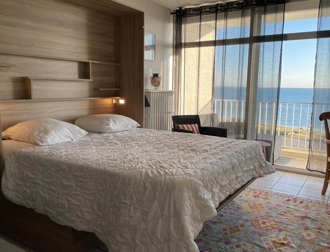 A spacious bed is incorporated into a wooden wall unit, complemented by soft bedding. Large glass doors provide access to a balcony, revealing views of the sea. Natural light floods the room through sheer black curtains, while a small seating area is set nearby.