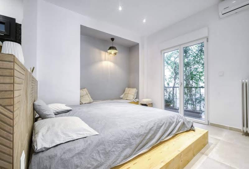 A spacious sleeping area features a low wooden platform bed with neutral bedding. Soft armchairs are positioned beside a large window that opens to a balcony, showcasing greenery outside. Recessed lighting highlights the room's minimalist design and creates a calming environment.