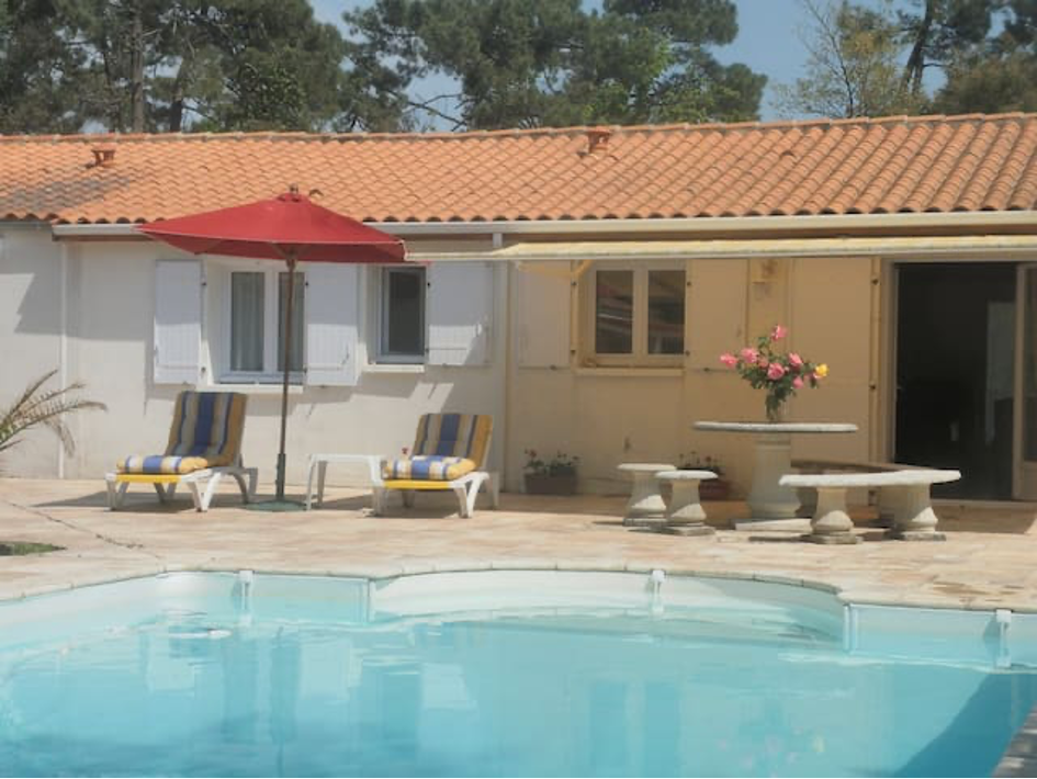 A tranquil outdoor space features a swimming pool surrounded by paved areas. Two striped lounge chairs sit next to the pool, while a round table with flowers is positioned nearby. An umbrella provides shade, and windows are visible on the house's façade.