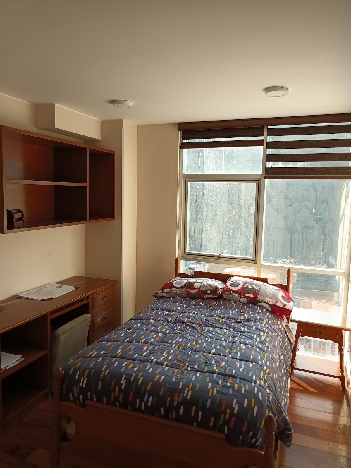 A well-lit bedroom features a spacious bed dressed with a patterned quilt. A wooden desk with drawers and a chair is positioned nearby. Natural light enters the room through a large window adorned with striped shades, creating a bright and functional space.
