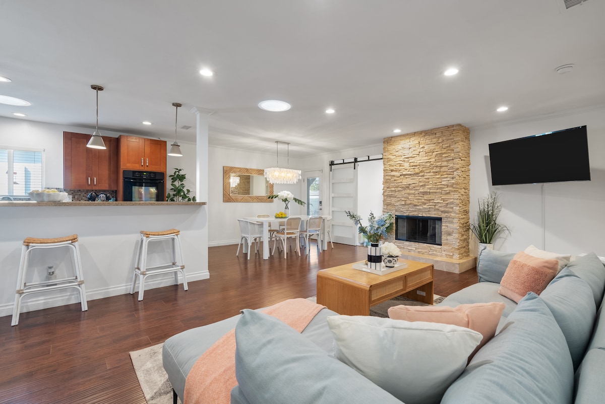 An open living space is bordered by a stylish kitchen and dining area. A comfortable sectional sofa is adorned with pastel cushions, facing a stone fireplace. Natural light fills the room, highlighting a dining table surrounded by chairs and a large wall-mounted TV.