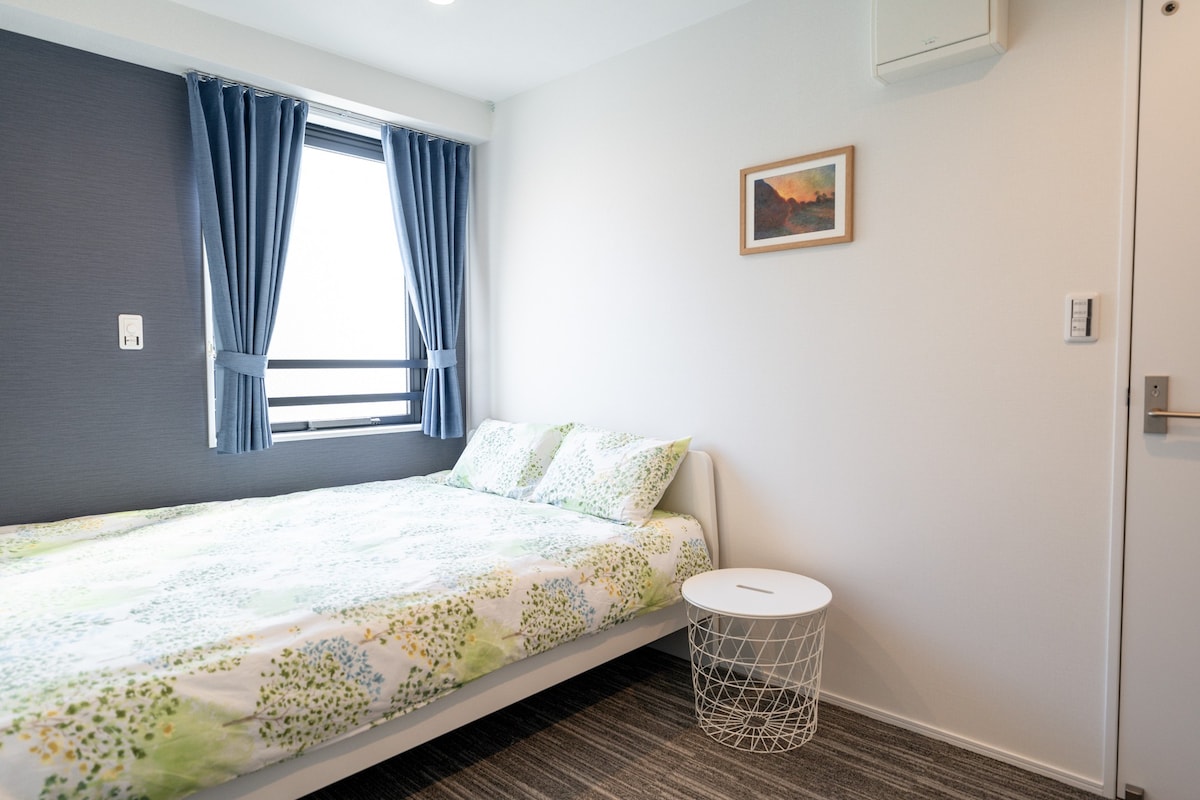A bright sleeping area is showcased, featuring a spacious bed adorned with a green-patterned spread. Natural light enters through a window dressed with blue curtains. A small white side table adds a functional touch next to the bed, while a framed art piece decorates the wall.