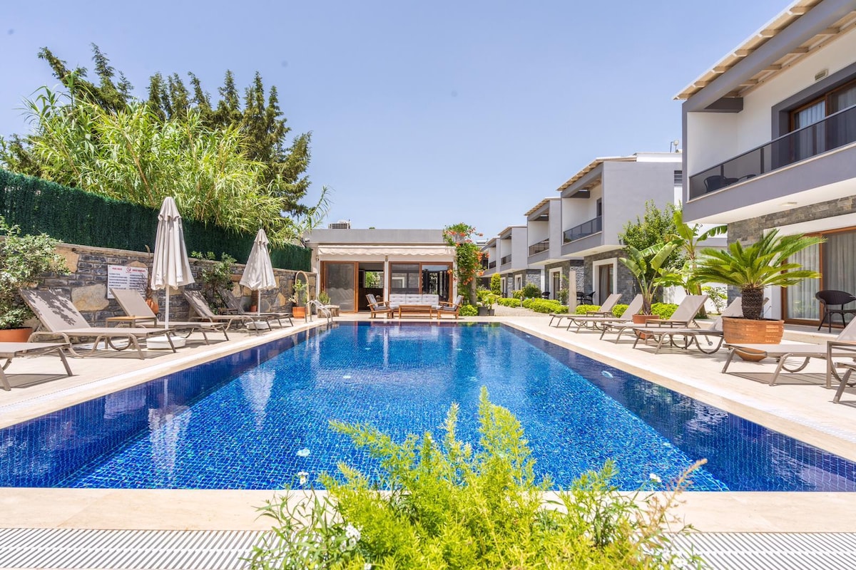 A tranquil pool area is surrounded by sun loungers and lush greenery. Four umbrellas provide shade near the pool's edge, while a wooden structure offers a shaded seating area. The buildings in the background feature balconies overlooking the pool.