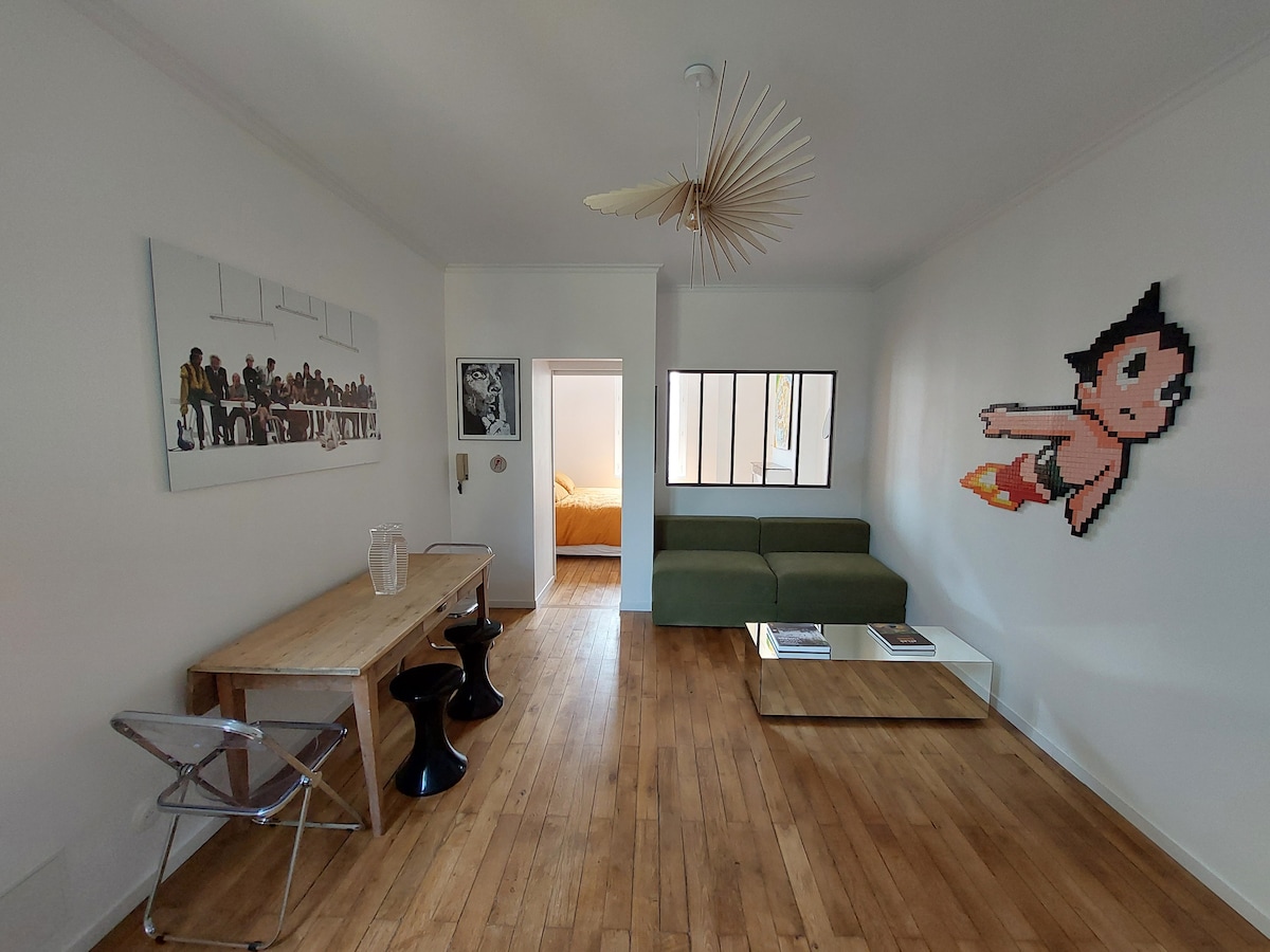 A living area features a green sofa, complemented by a wooden table and black stools. A large artwork is displayed on one wall, while a colorful character mural adds a playful touch. Light streams through a window, illuminating the wood flooring.