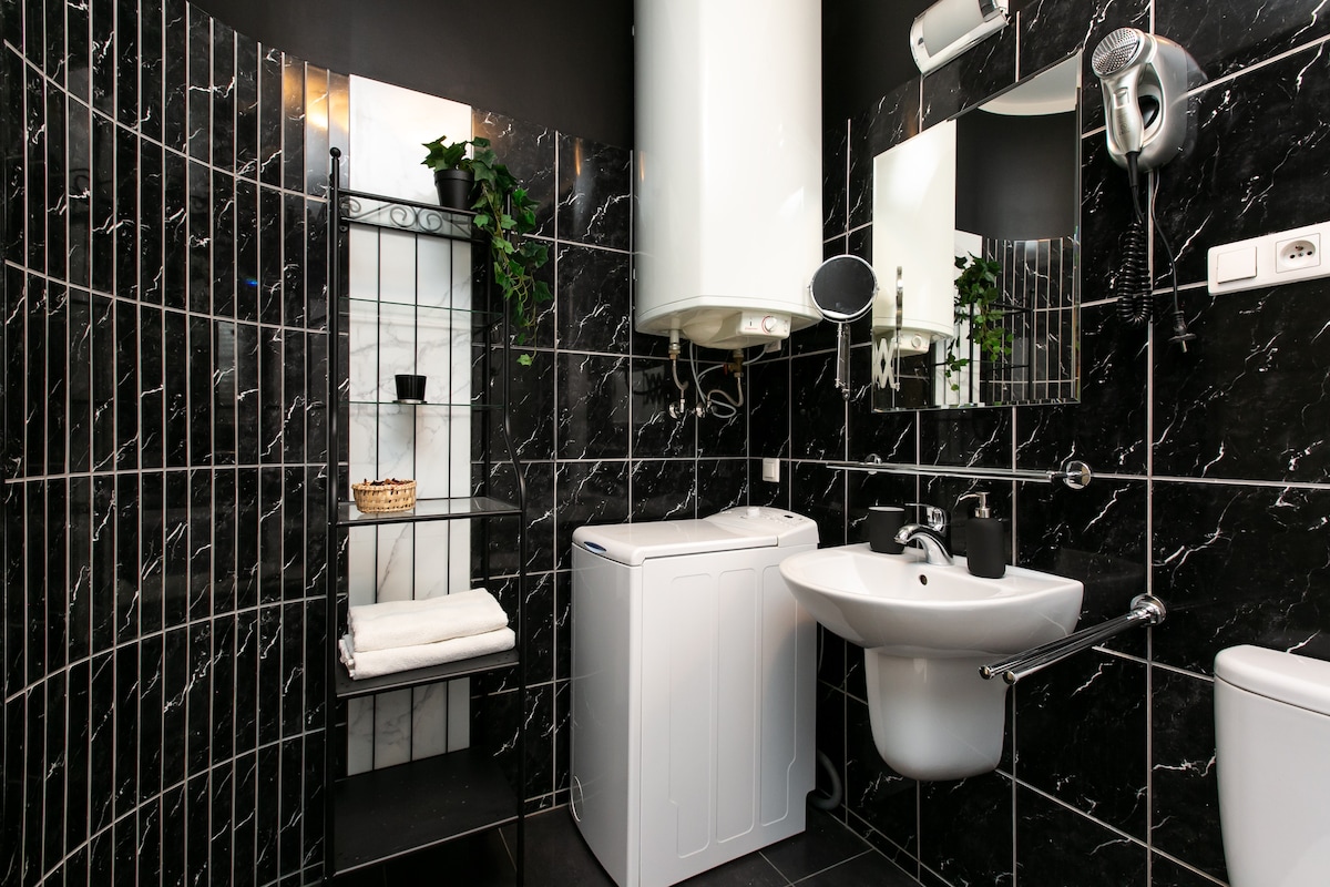 A modern bathroom features dark patterned tiles along the walls and floor. A compact washing machine is placed beside a sleek washbasin, and a mirror is mounted above it. Shelving holds neatly stacked towels and small plants add a touch of greenery.