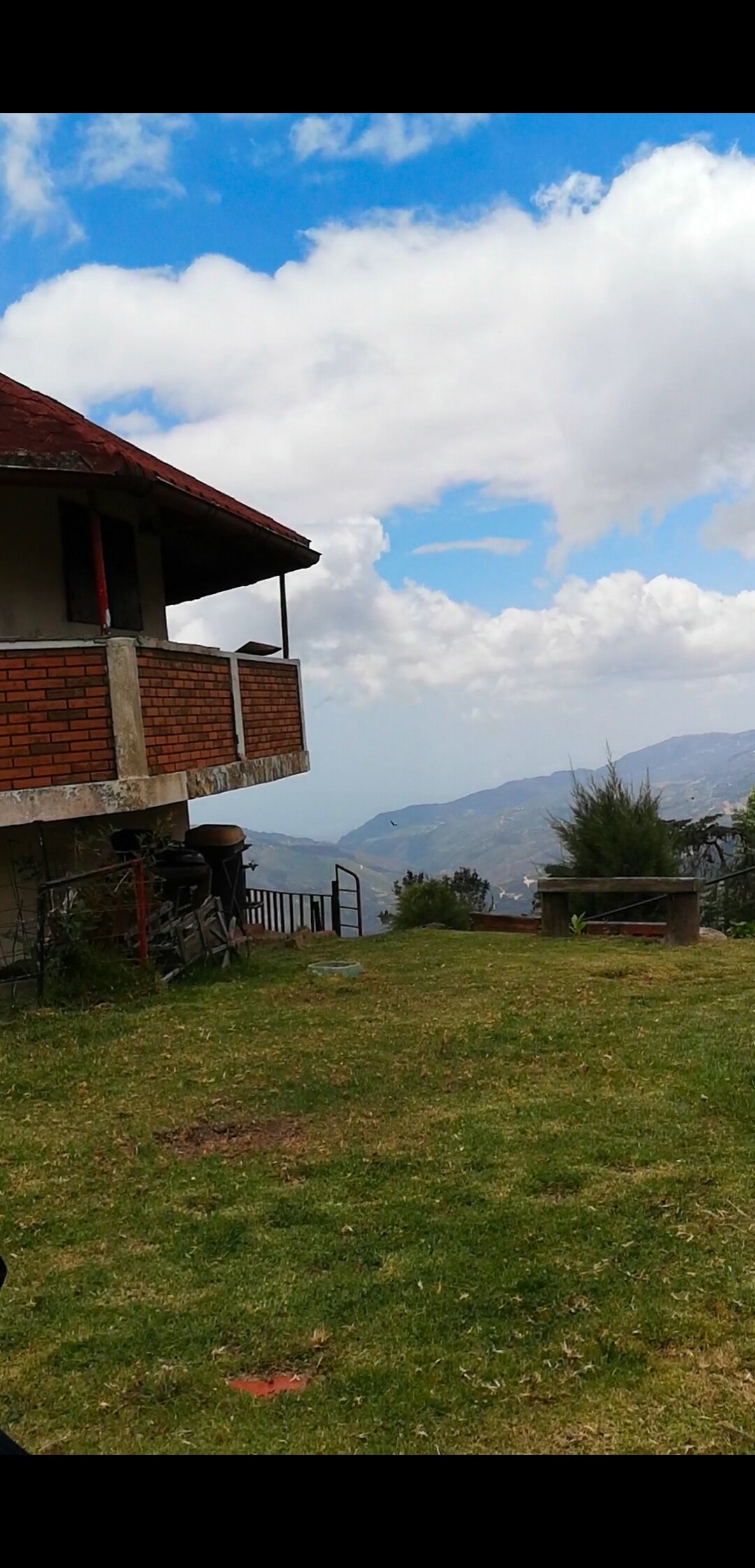 A two-story house with a reddish roof overlooks a green landscape. The balcony offers views of distant mountains under a partly cloudy sky. A small outdoor seating area is positioned on the grassy lawn, providing a peaceful spot to enjoy the surroundings.