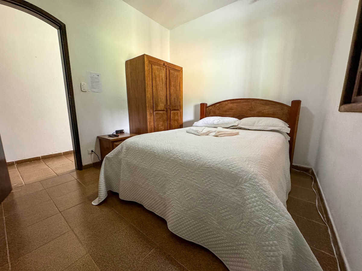 A cozy bedroom features a spacious bed covered with a light quilt. A rustic wooden closet stands against one wall, and a side table accompanies the bed. Soft natural light filters through the window, illuminating the neutral-colored tiled floor.