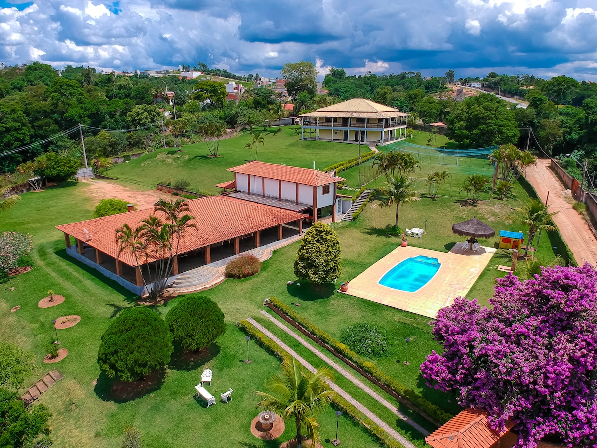 An aerial view presents a well-maintained property featuring a vibrant green lawn, surrounded by trees and colorful flowers. A swimming pool is visible beside a shaded seating area, with additional structures in the background, including the main building. The landscape is open and inviting.