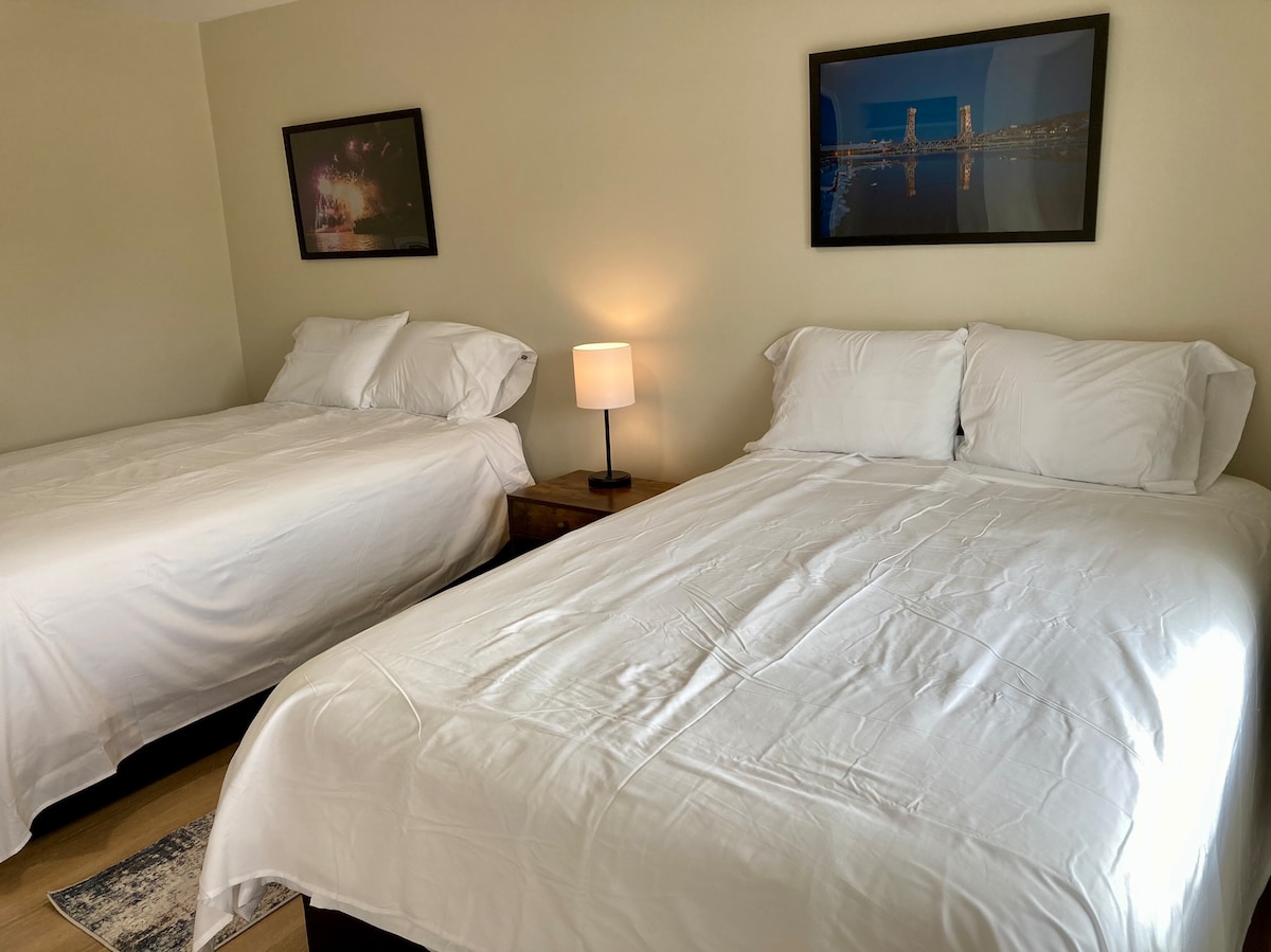 A spacious sleeping area features two large beds covered in white linens, creating a clean and inviting look. A bedside lamp provides soft illumination, while two framed photographs hang on the wall, adding subtle elegance to the room.