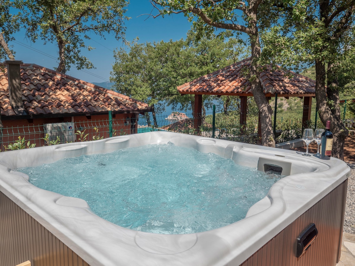 A private hot tub is situated outdoors, surrounded by greenery and offering serene views. The tub features a spacious design with bubbling water, complemented by a nearby shaded area with a tiled roof. A bottle of wine rests on the edge of the tub.