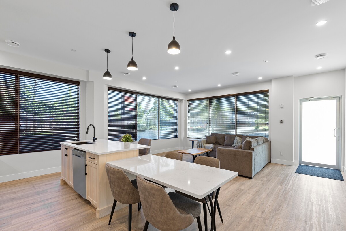 A modern open-concept living area features large windows with blinds, allowing natural light to filter in. A spacious kitchen island stands next to a dining table and four chairs, while a comfortable seating area with a sofa is arranged to foster relaxation.