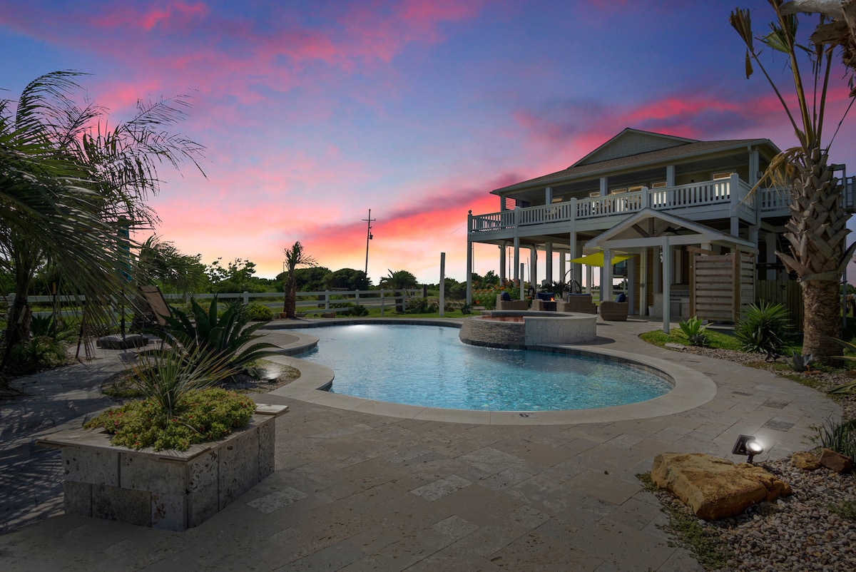 The image captures a private saltwater pool surrounded by tropical landscaping. The pool features a hot tub with a cascading water feature. A two-story home stands in the background, with a large covered deck offering seating and ocean views, enhanced by a colorful sunset sky.
