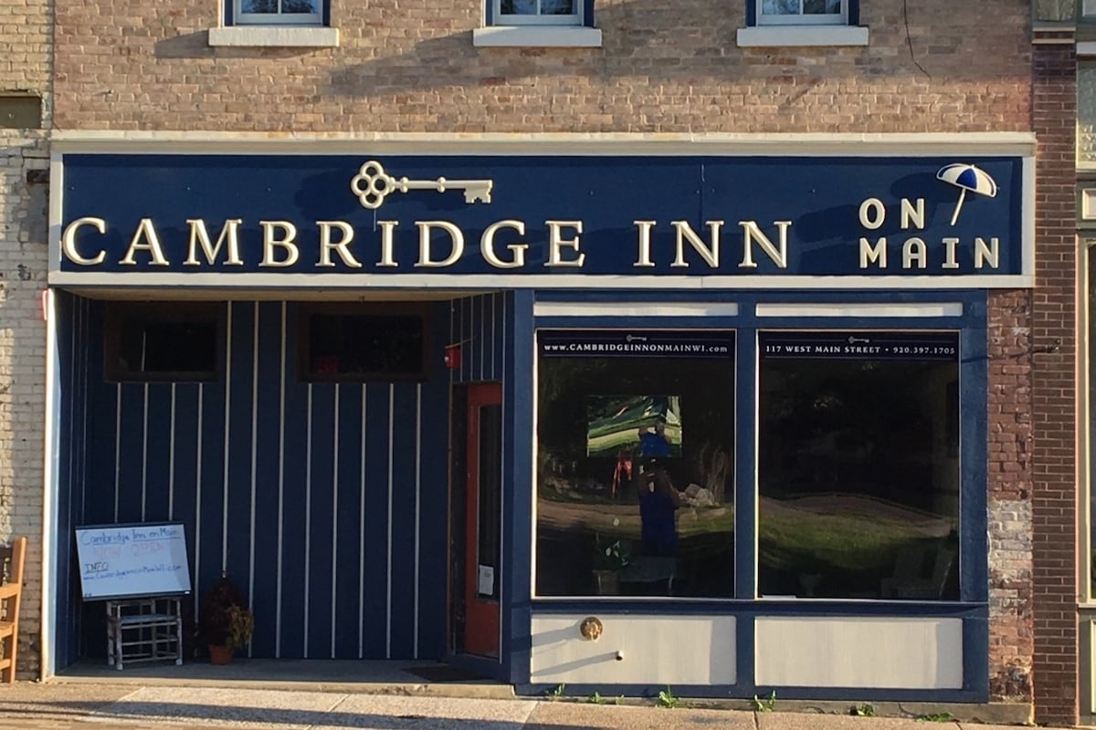 The exterior of the Cambridge Inn features a charming blue and white color scheme with large windows reflecting the surroundings. A decorative key and umbrella signage enhances the entrance. The sidewalk showcases a small seating area, providing a warm welcome to guests.