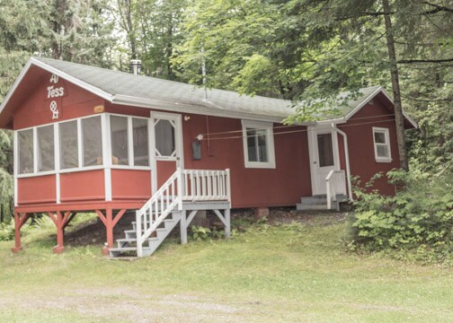 A red chalet is nestled among lush green trees, featuring a welcoming front porch with a screened-in area. The entrance is accessed via a set of steps, and the surrounding landscape includes dense foliage and natural scenery.