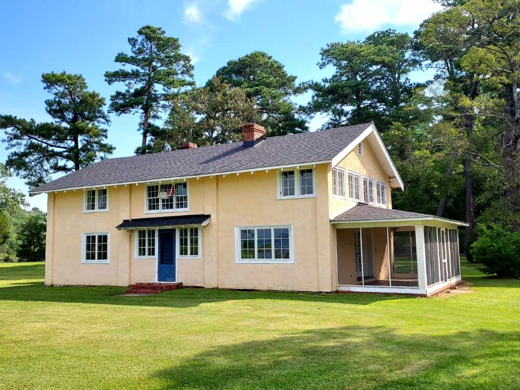 The exterior of the converted Log Cabin School is showcased, revealing a two-story structure with a beige facade and white trim. A spacious screened porch extends from one side. Lush green lawns surround the home, while tall trees provide shade and contrast against the clear blue sky.