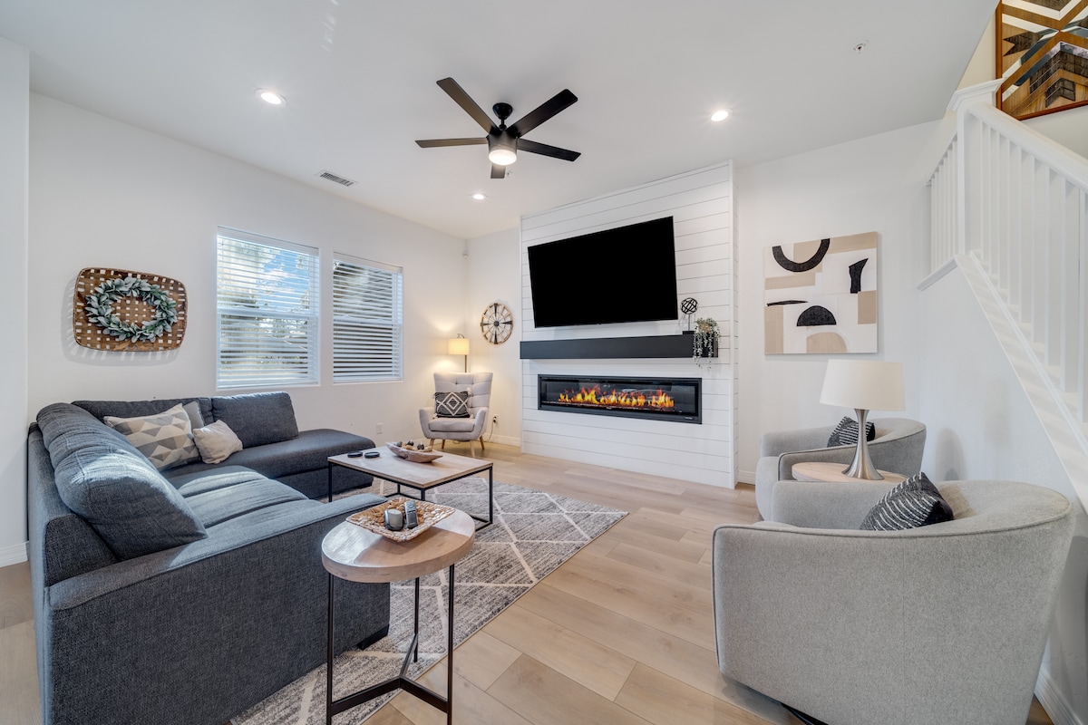 A cozy living area features a large sectional sofa complemented by plush chairs. A modern fireplace is installed beneath a flat-screen TV, providing a focal point. Natural light enters through multiple windows, and a stylish area rug enhances the space.