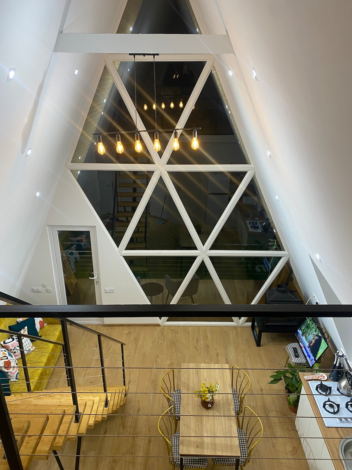 An airy interior showcases a diagonal wall of large triangular windows that allow natural light to fill the space. A dining area features a simple table adorned with a floral centerpiece, while a staircase leads to an upper level, enhancing the open layout.