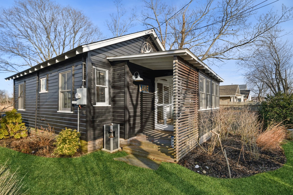 The exterior of a modern cottage is presented with a black wood facade and multiple windows. A front porch is seen, partially enclosed with slatted wood. Lush green grass and shrubbery frame the entrance, creating a welcoming approach to the home.