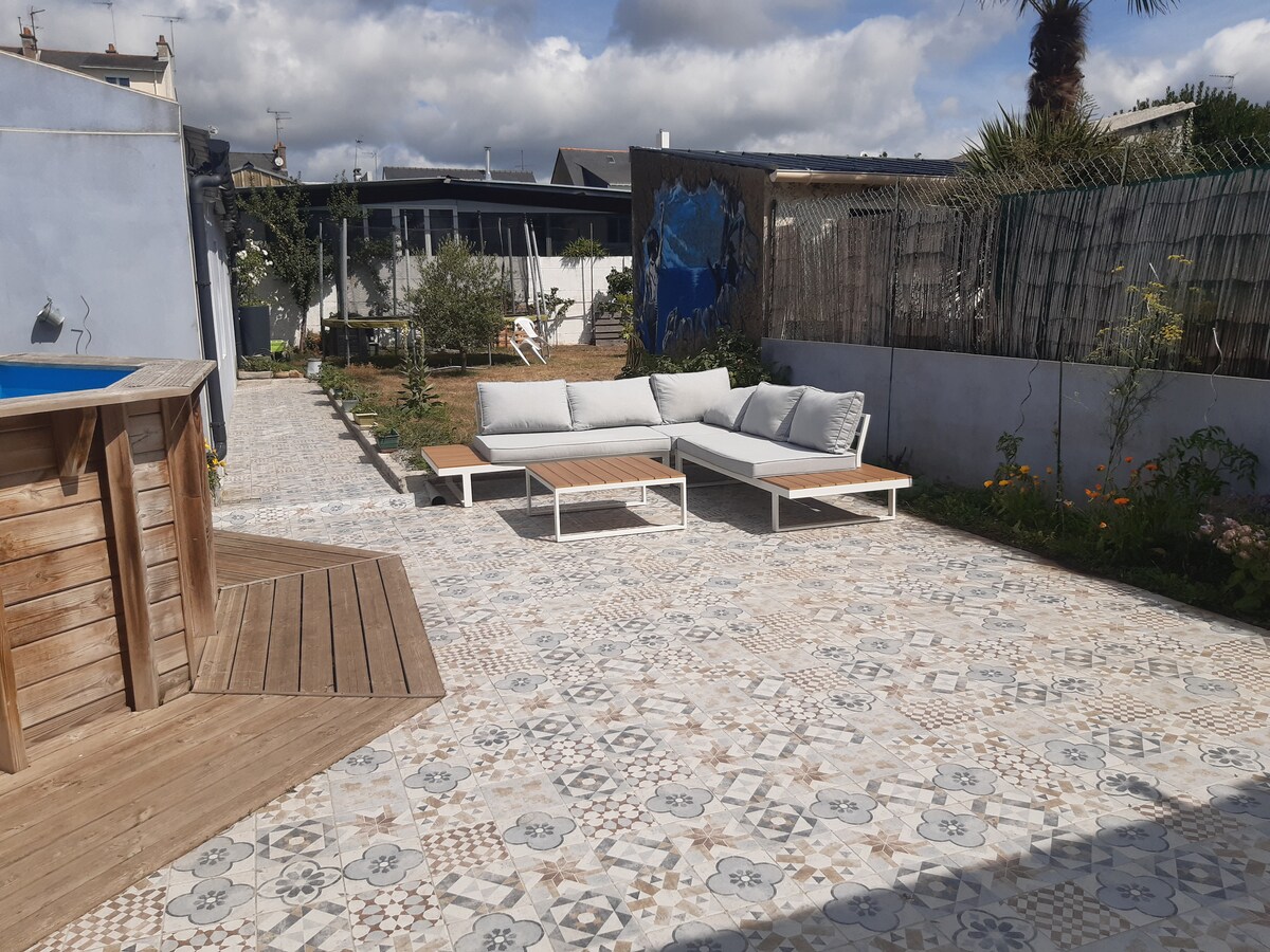 An outdoor space features a tiled patio with a seating area consisting of light-colored couches and a wooden table. A small pool can be seen to the left, surrounded by greenery. The sky is partly cloudy, adding depth to the serene environment.
