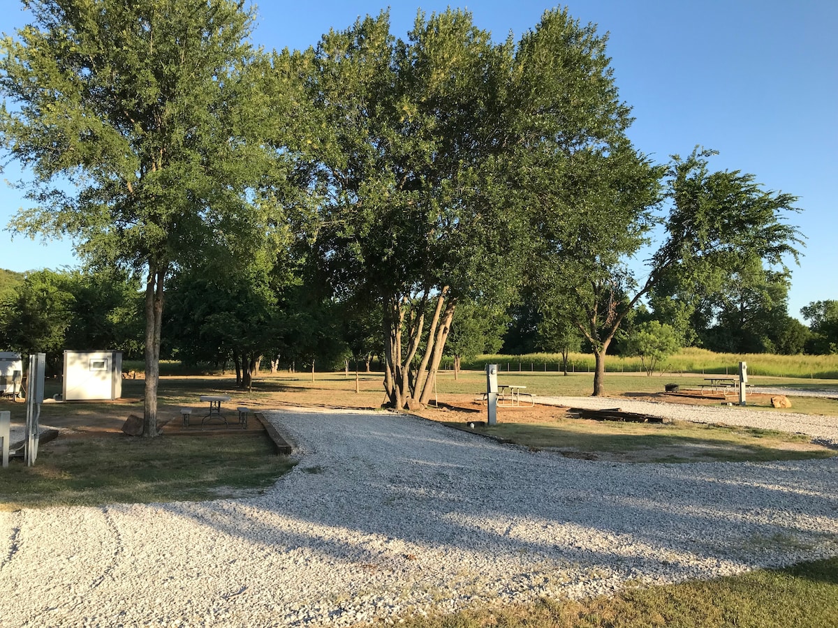 A gravel area is visible, featuring several shaded spots created by large trees. Two picnic tables are positioned nearby, while utility hookups can be seen at designated spots. The open space offers a natural setting with the backdrop of a green landscape.
