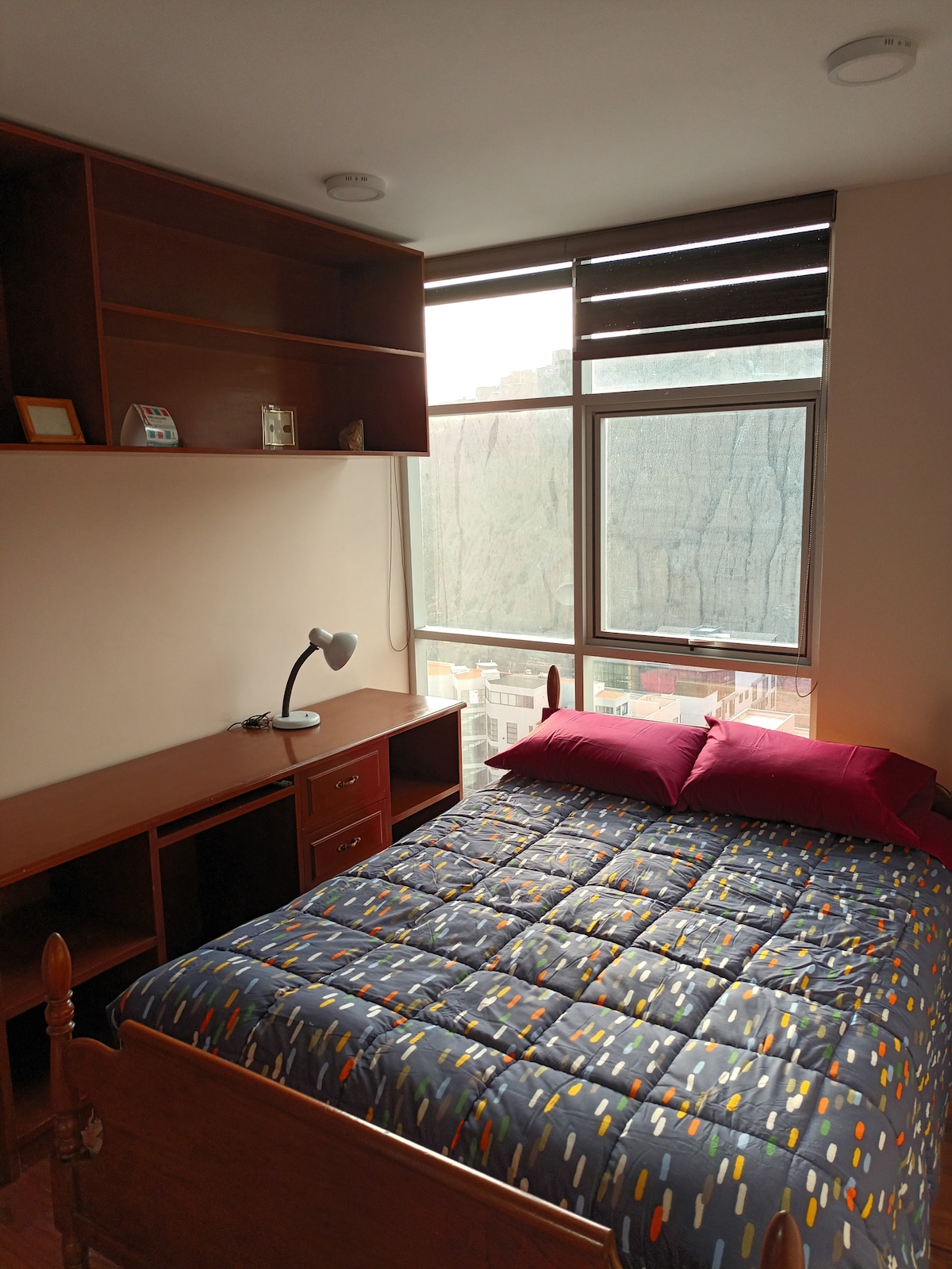 A cozy bedroom features a spacious bed with a patterned comforter and pink accent pillows. A wooden desk with a lamp is positioned near a large window, providing natural light and views of the surrounding landscape. A shelf is mounted above the desk for additional storage.