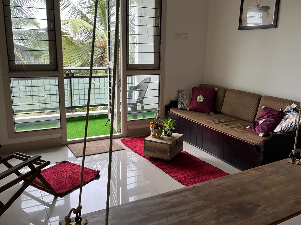 A living area features a comfortable sofa adorned with patterned cushions, complemented by a rustic coffee table. Light floods through large glass doors, surrounded by lush greenery visible outside. A swing chair adds unique charm to the space, enhancing relaxation.
